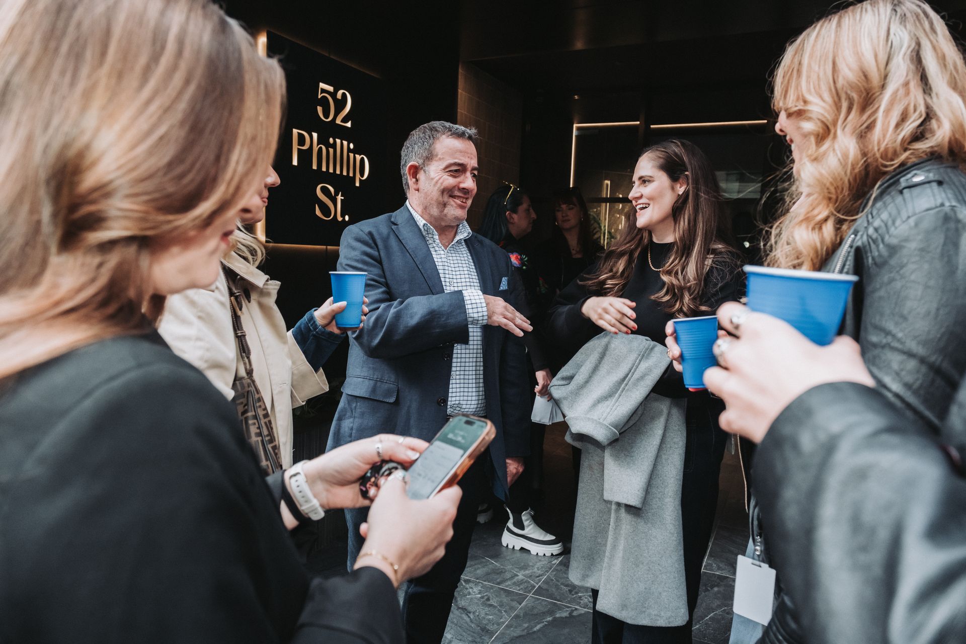 People outside a building at 52 Phillip St, some holding blue cups, chatting.