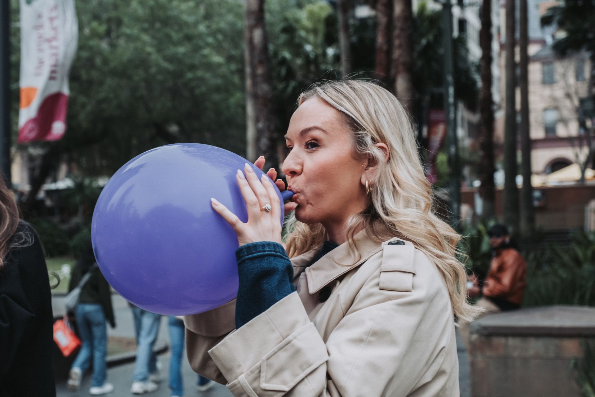 Woman blowing up a large purple balloon outdoors. She wears a tan coat and has blonde hair.
