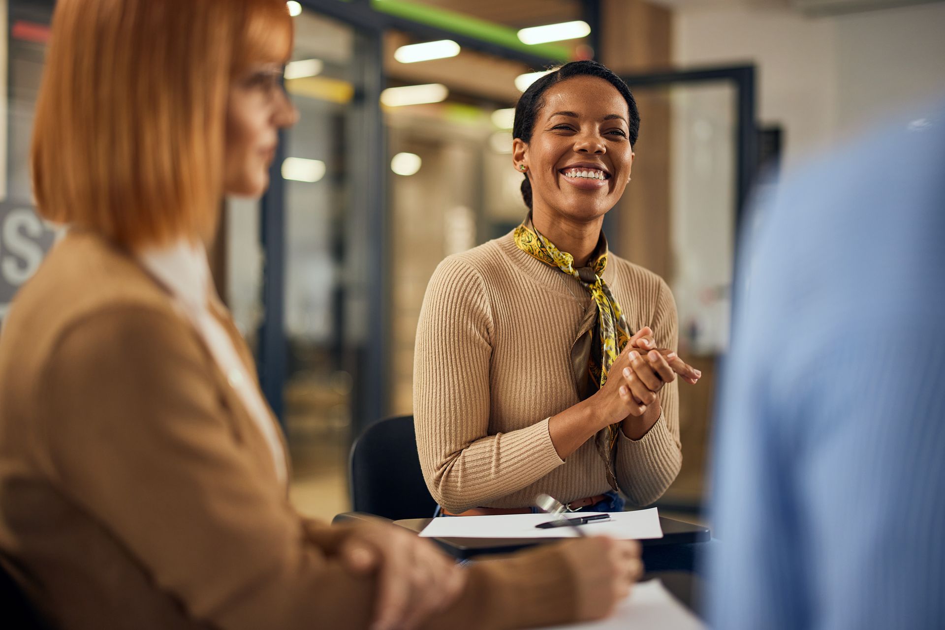 Woman smiling during a meeting, hands clasped. Two other people are partially visible in office setting.