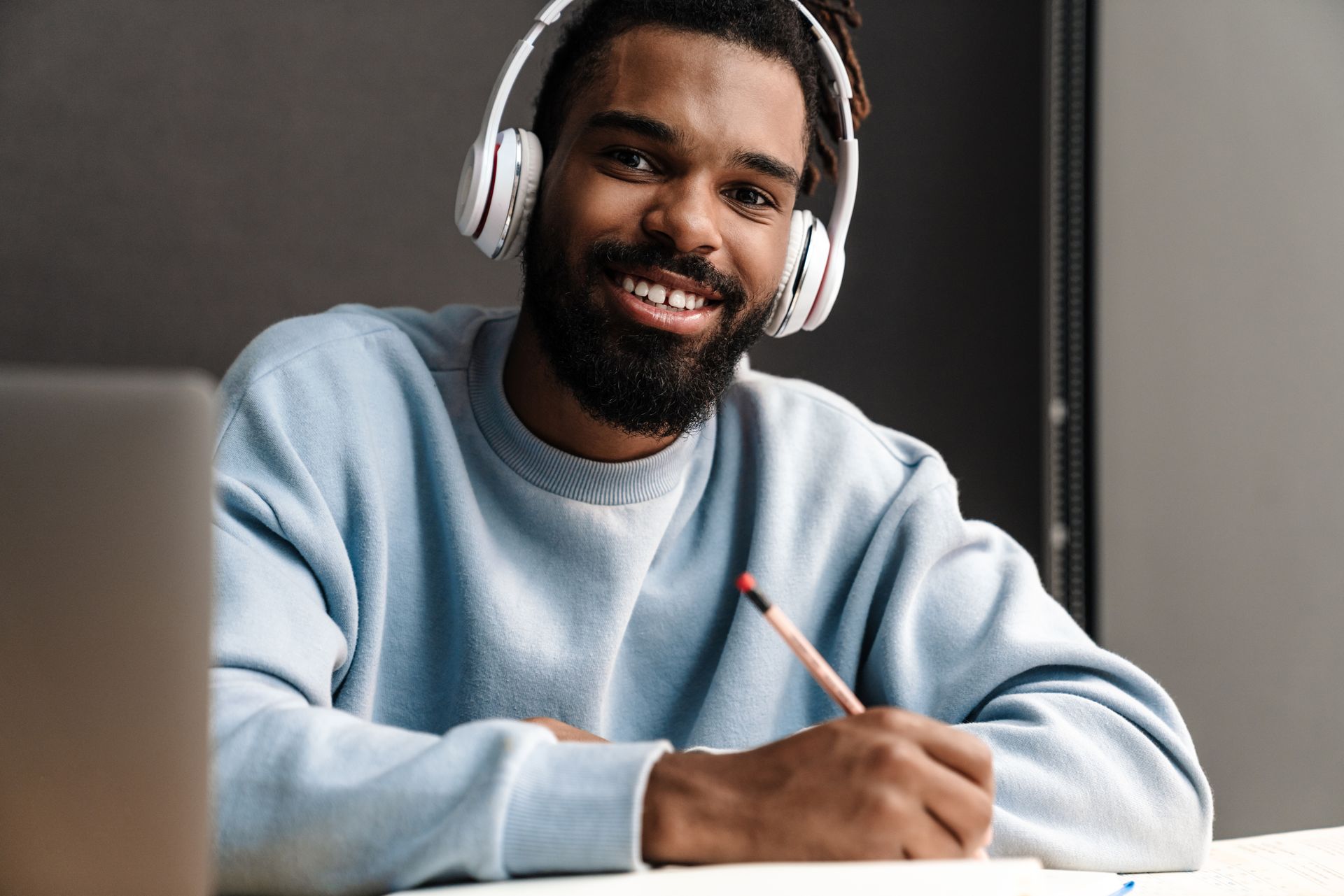 Man wearing headphones smiles while writing at a desk with a laptop.