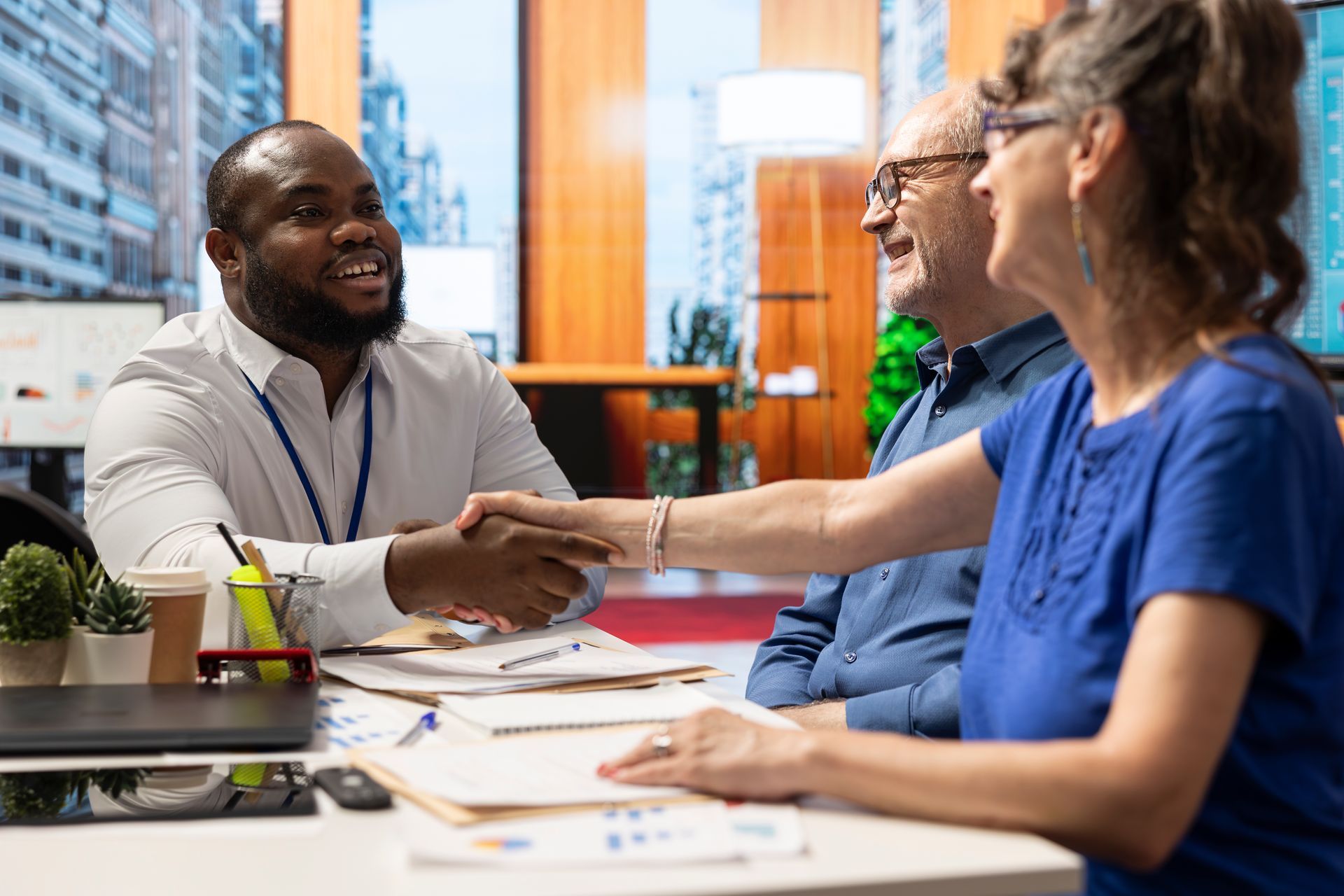 Man shaking hands with a person at a table, smiling. Two other people are in the background, in an office.