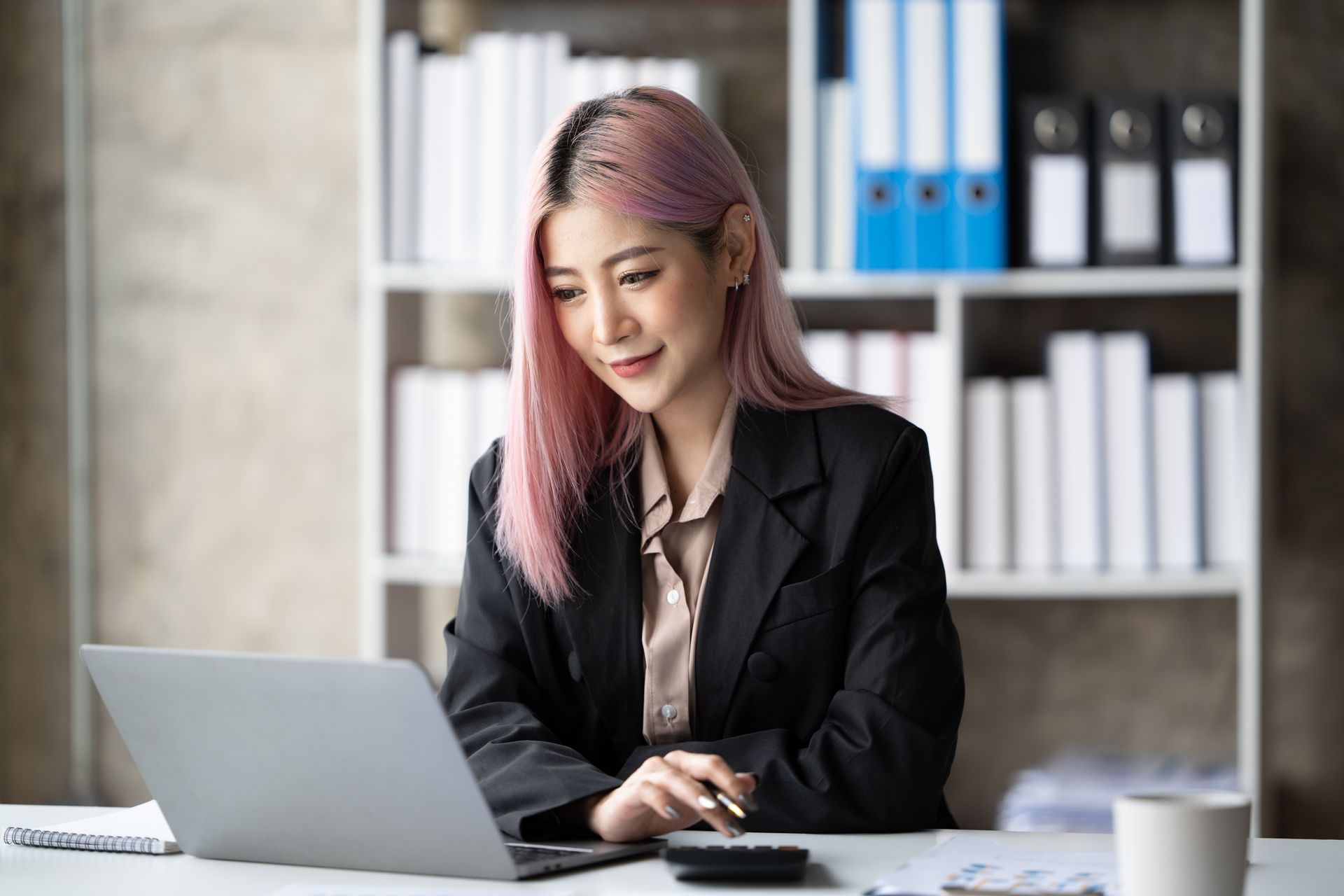 Woman with pink hair in a blazer working on a laptop at a desk with a calculator. Shelf in the background.