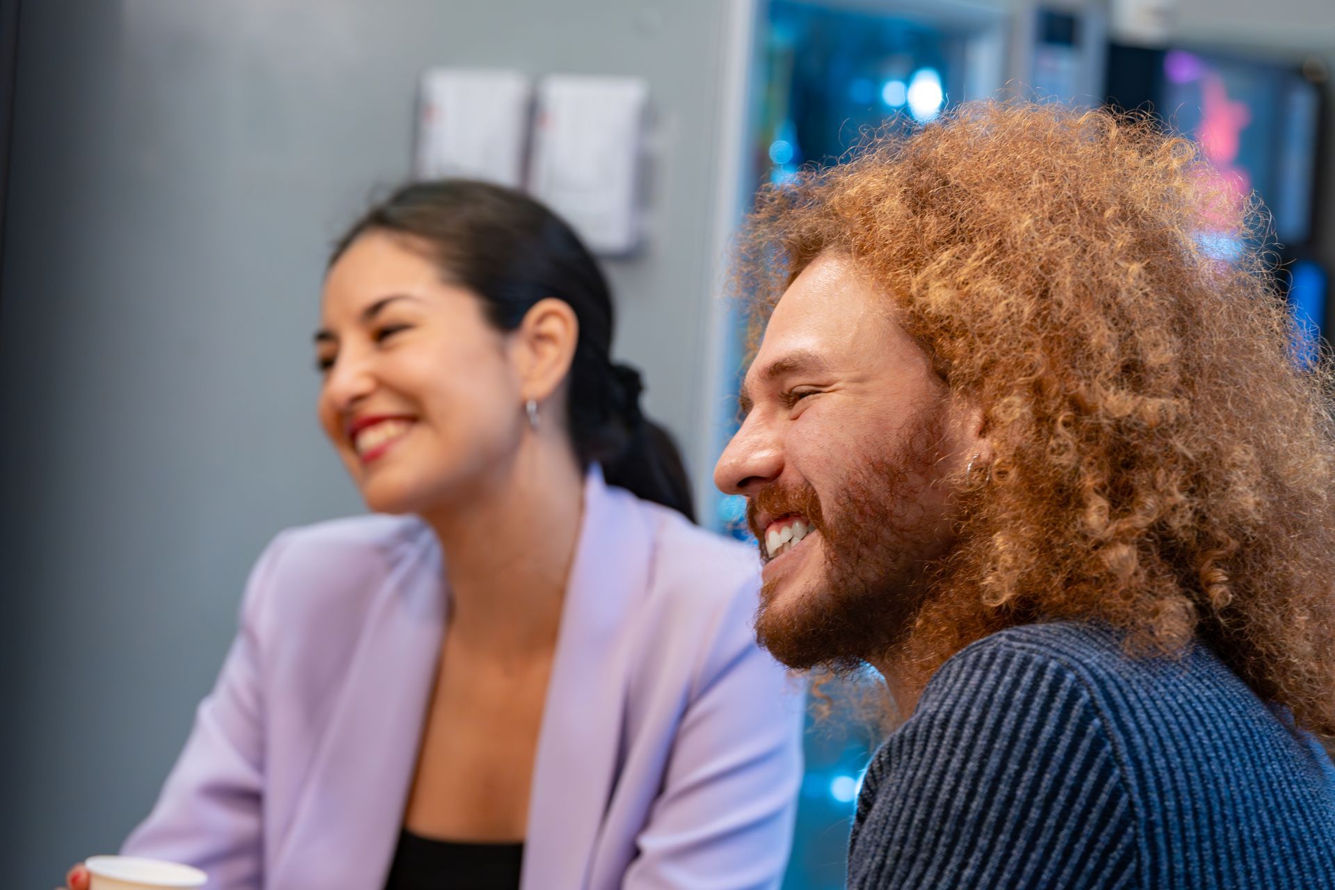 Woman in lilac jacket and man with red curly hair smile, sitting indoors.