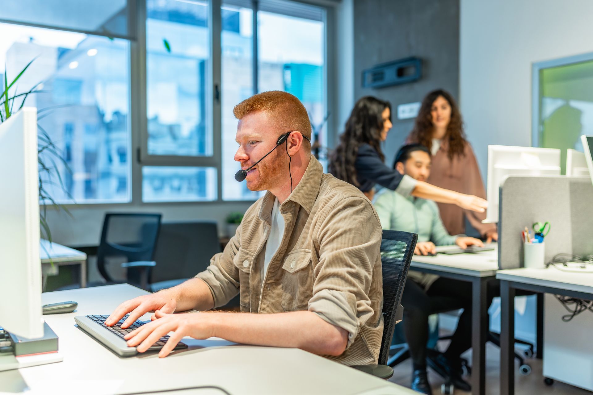 Man with headset types at computer; coworkers collaborate in a modern office setting.