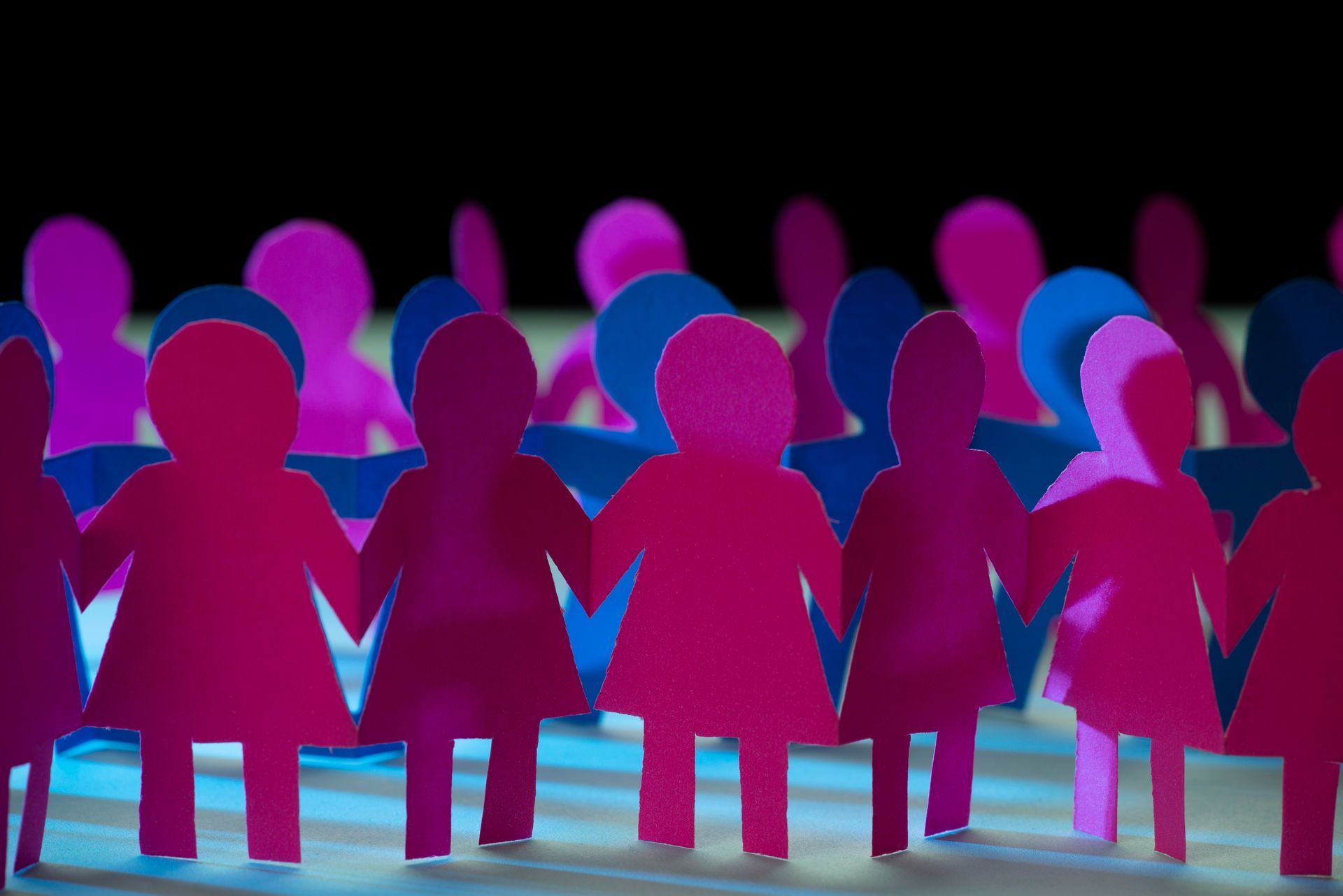 Pink and blue paper dolls holding hands, arranged in a chain against a dark background.