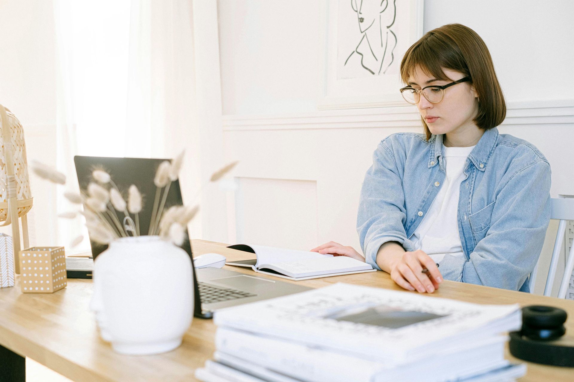 Woman wearing glasses looks down at a notebook at a desk with a laptop and a vase of flowers.
