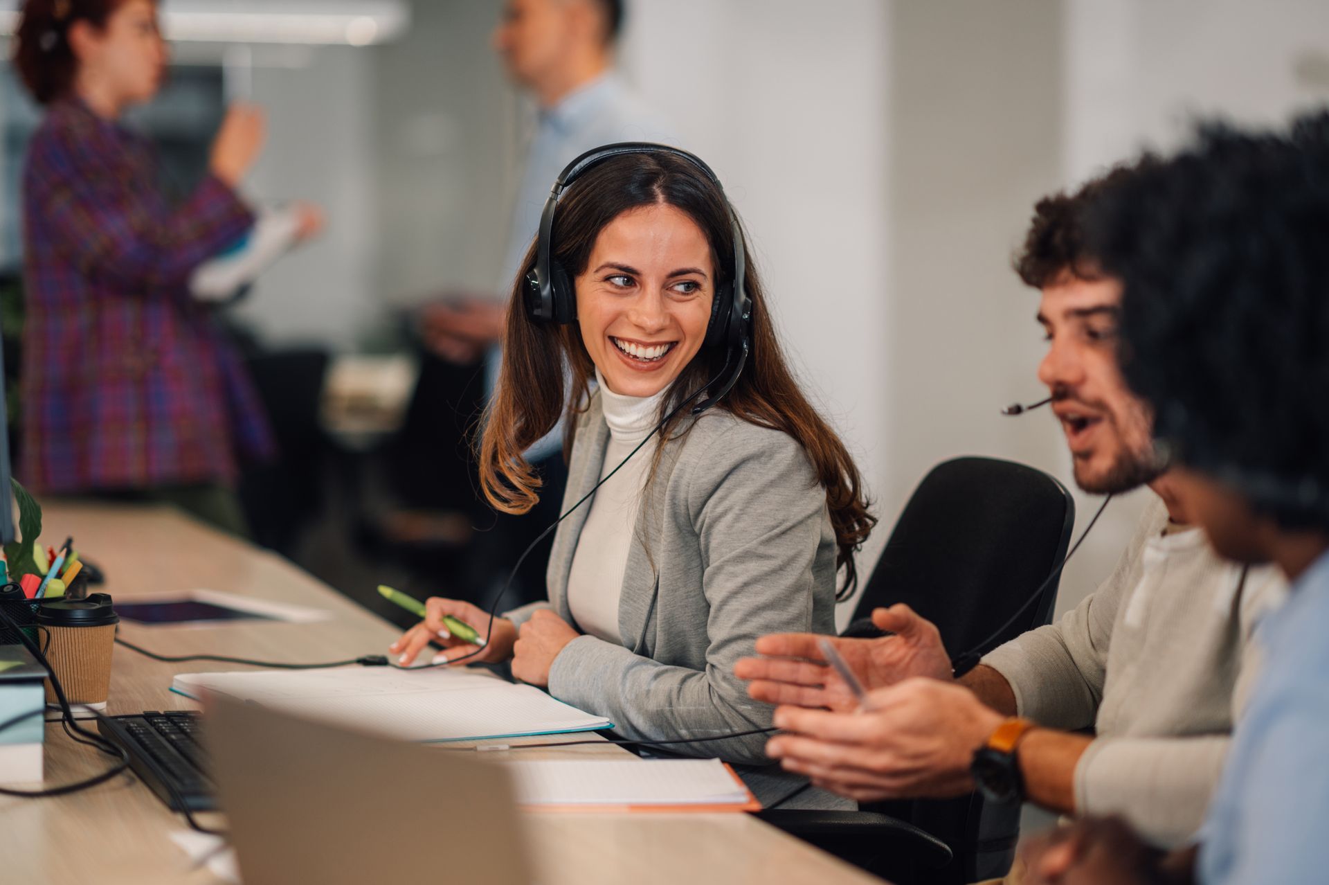 Office workers in conversation, woman with headset smiles, one gestures with hand.