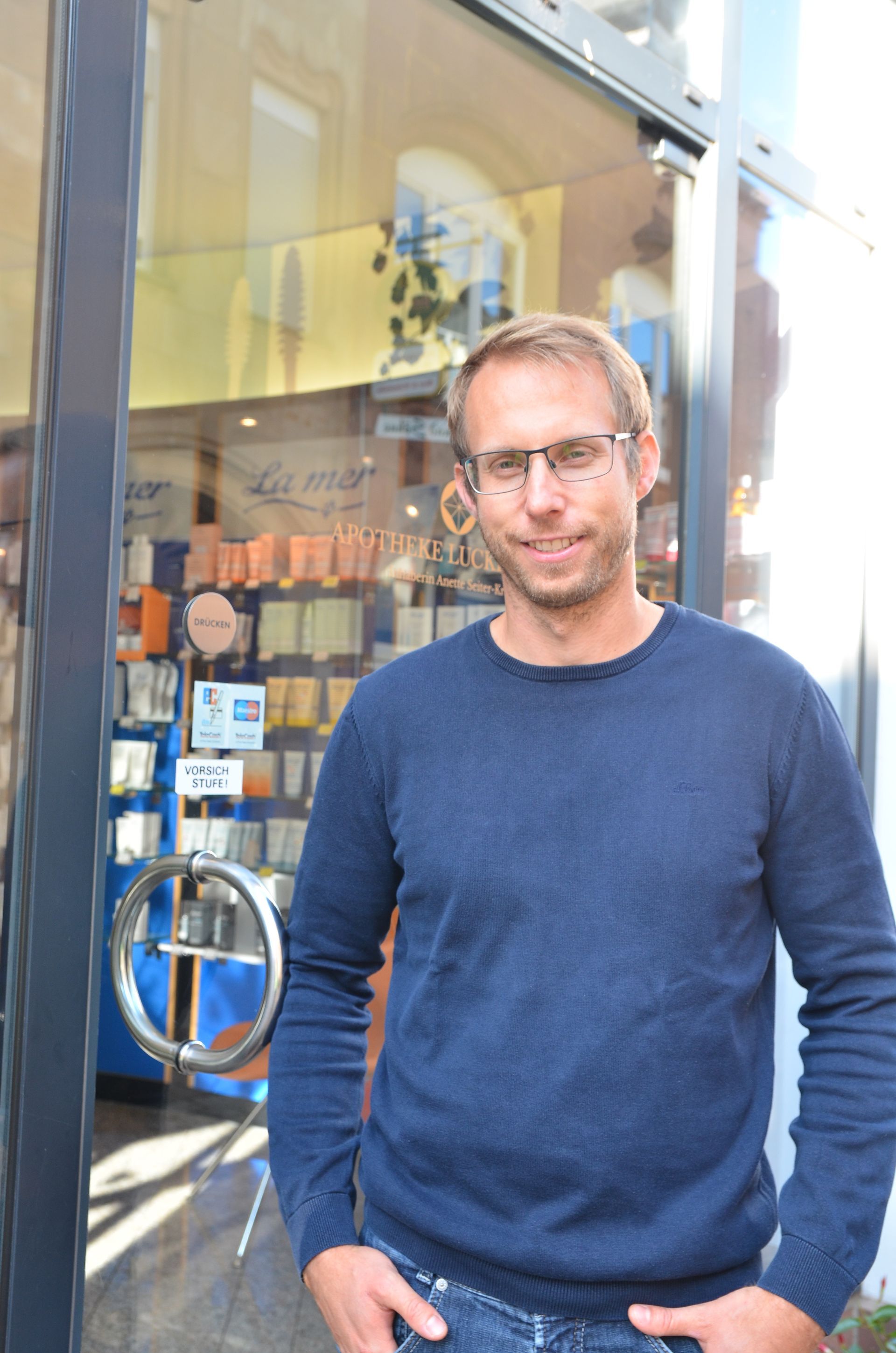 Ein Mann mit Brille und blauem Pullover steht vor einer Glastür.