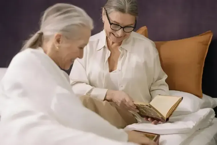 Two women in white sit on a bed, reading an open book together.
