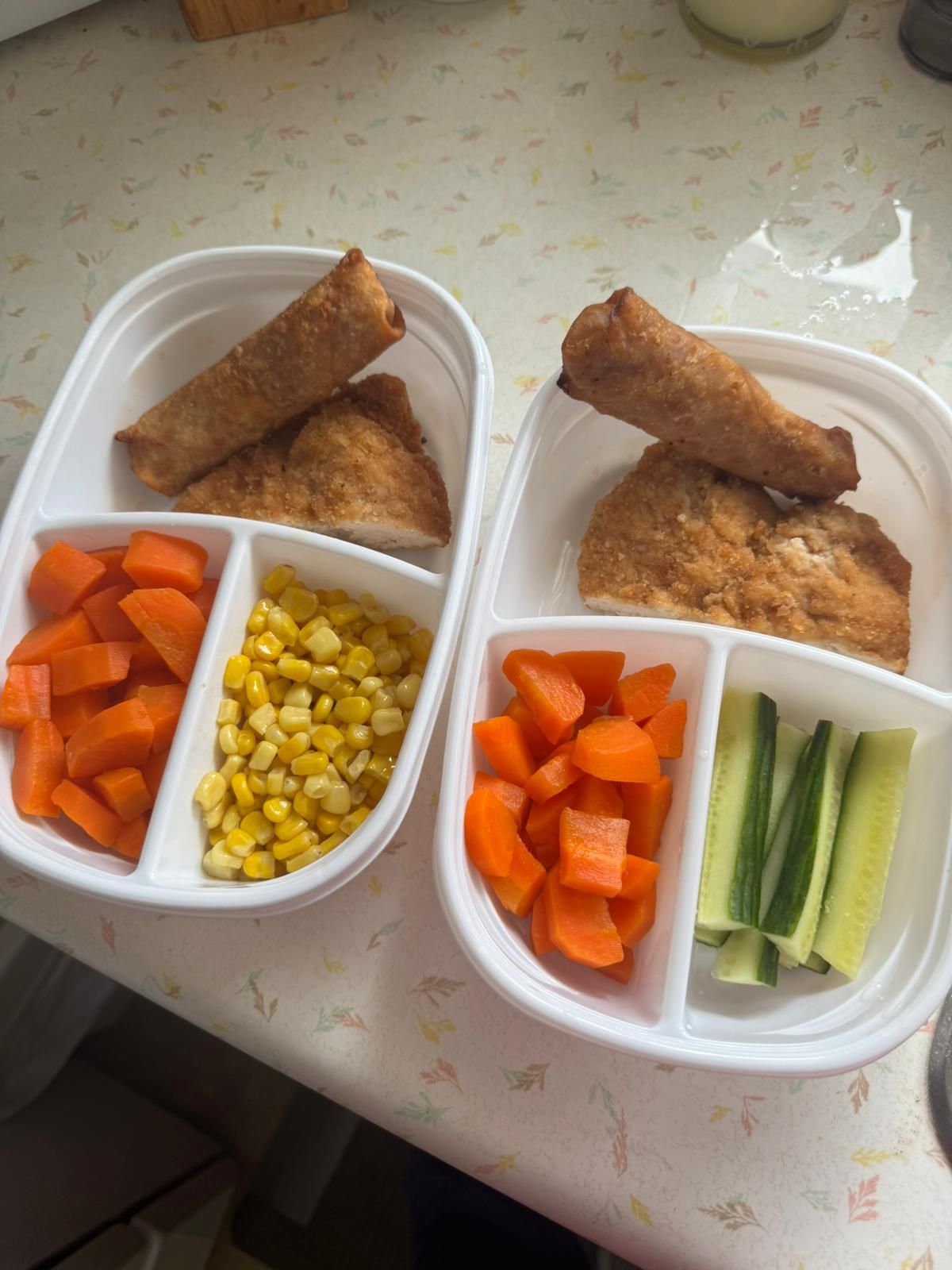 Two meal prep containers with fried rolls, carrots, corn, and cucumber sticks on a table