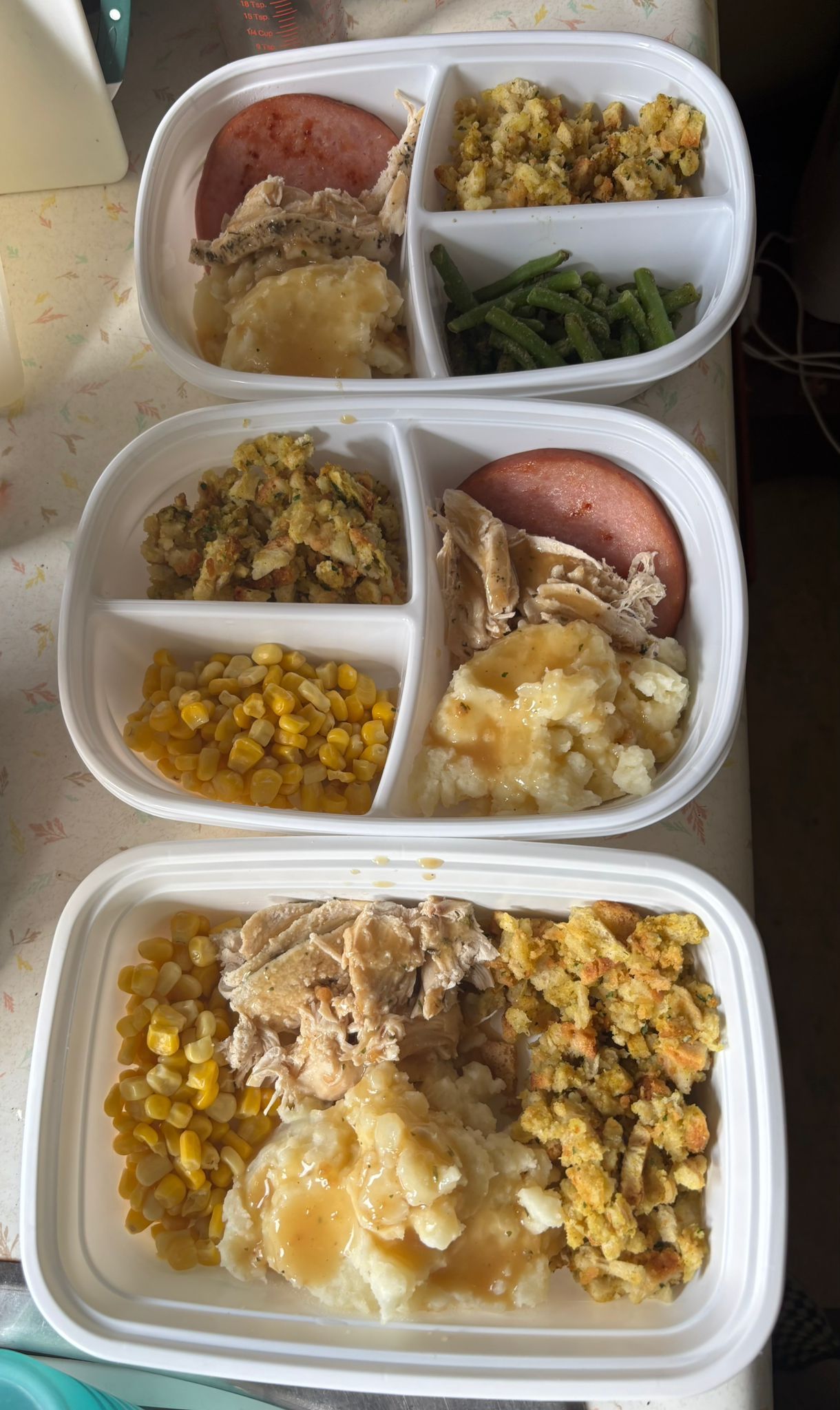Three meal-prep containers with corn, mashed potatoes, greens, and sliced meat in a fridge.
