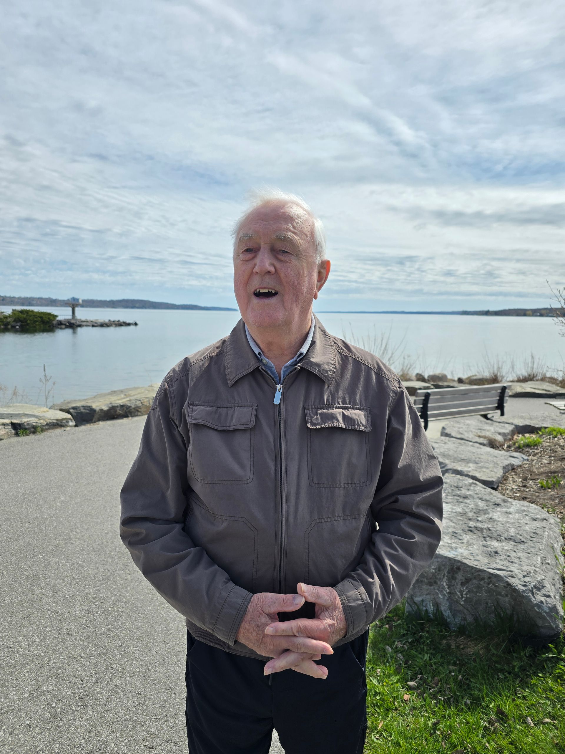 Man standing on a rocky shoreline by calm water under a cloudy sky, wearing a gray jacket.