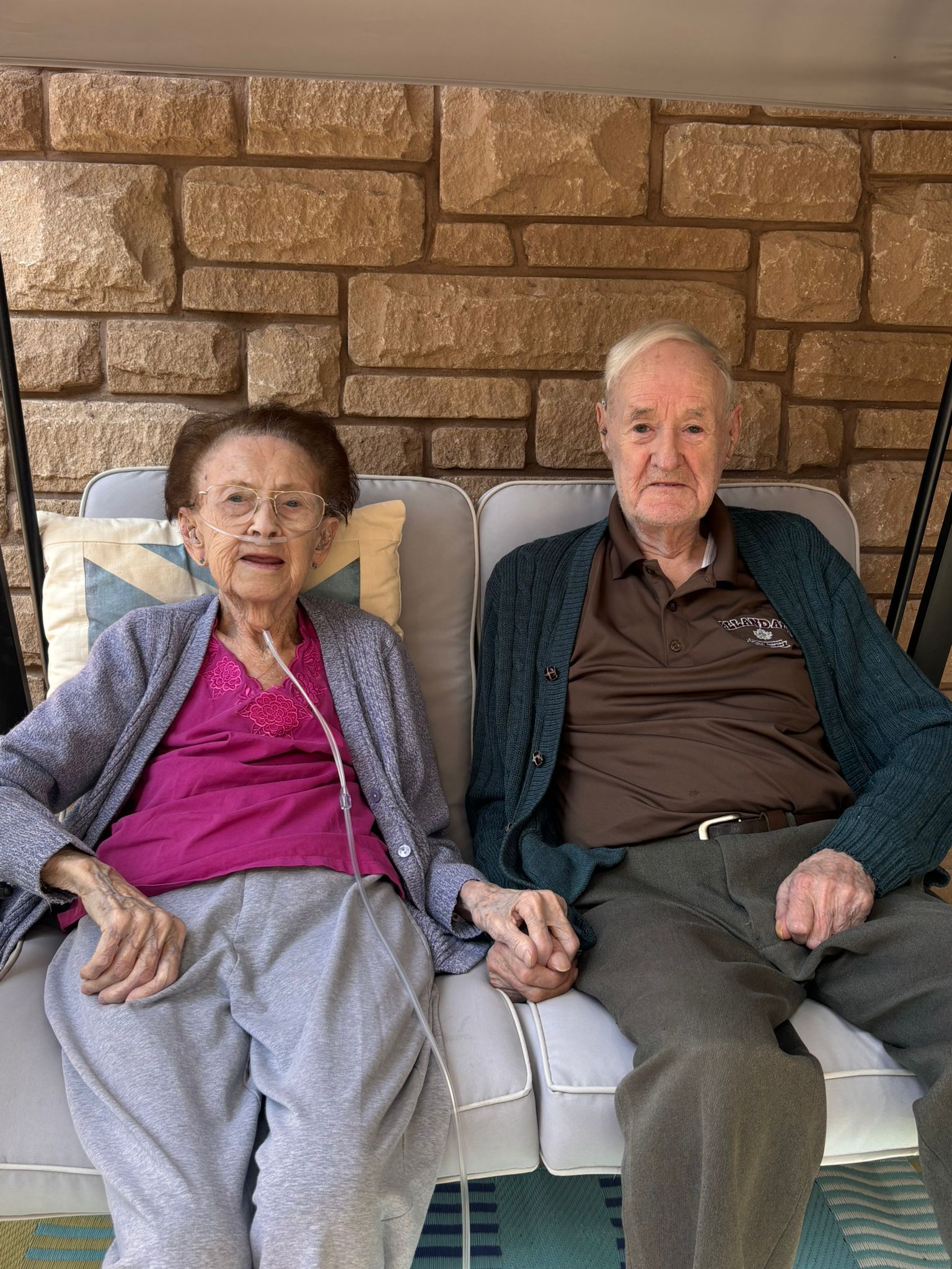 Two people sitting on a white couch in front of a stone wall, holding hands.