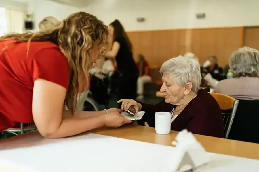 Woman in red shirt leaning toward seated older woman across a table in a community room