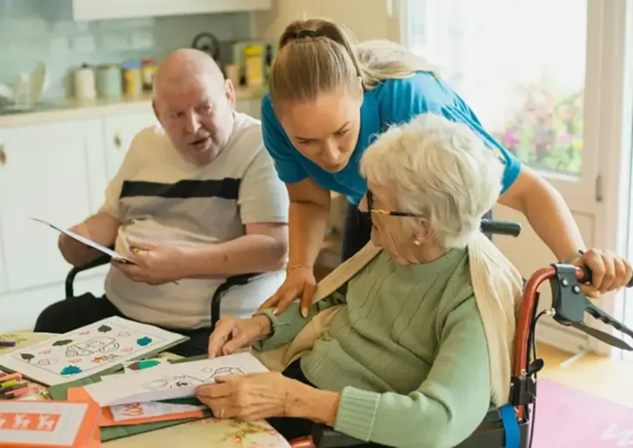Three people playing a board game at a table, with one person pointing and another assisting an elderly player.
