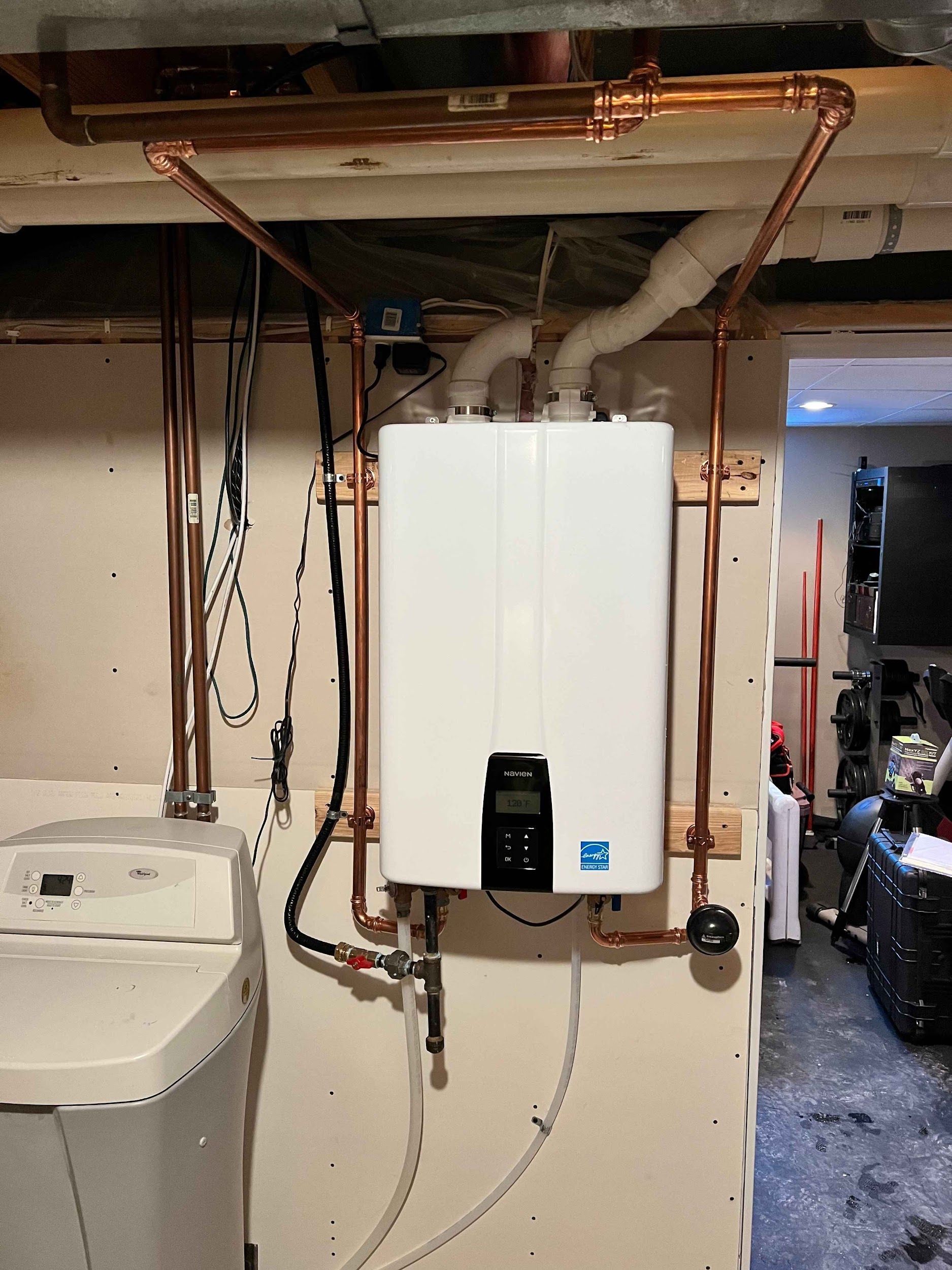 A white wall-mounted tankless water heater with copper pipes in a basement near a washing machine.