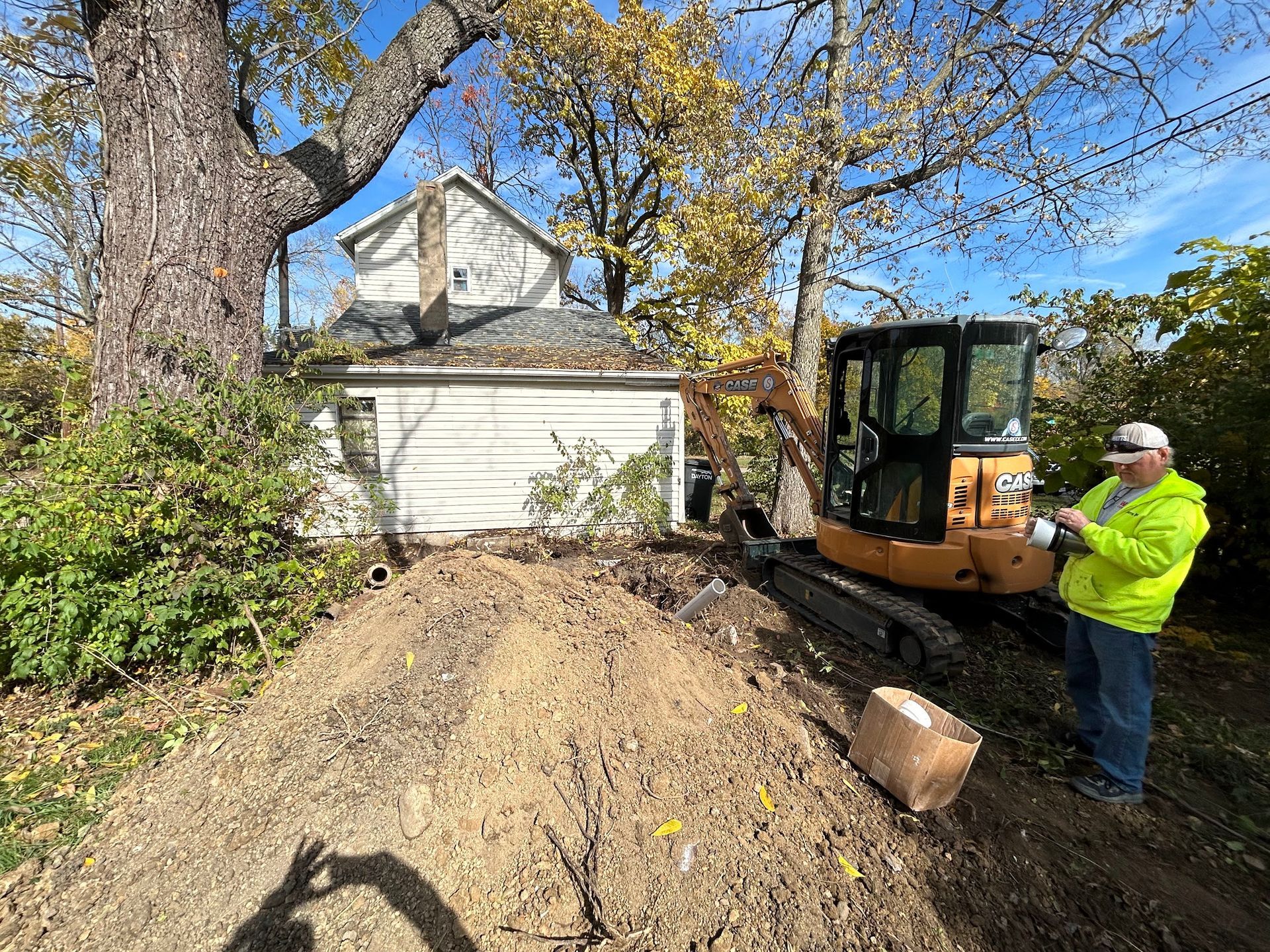 Man in neon vest operates excavator near a small white garage. Dirt pile in foreground, fall foliage.