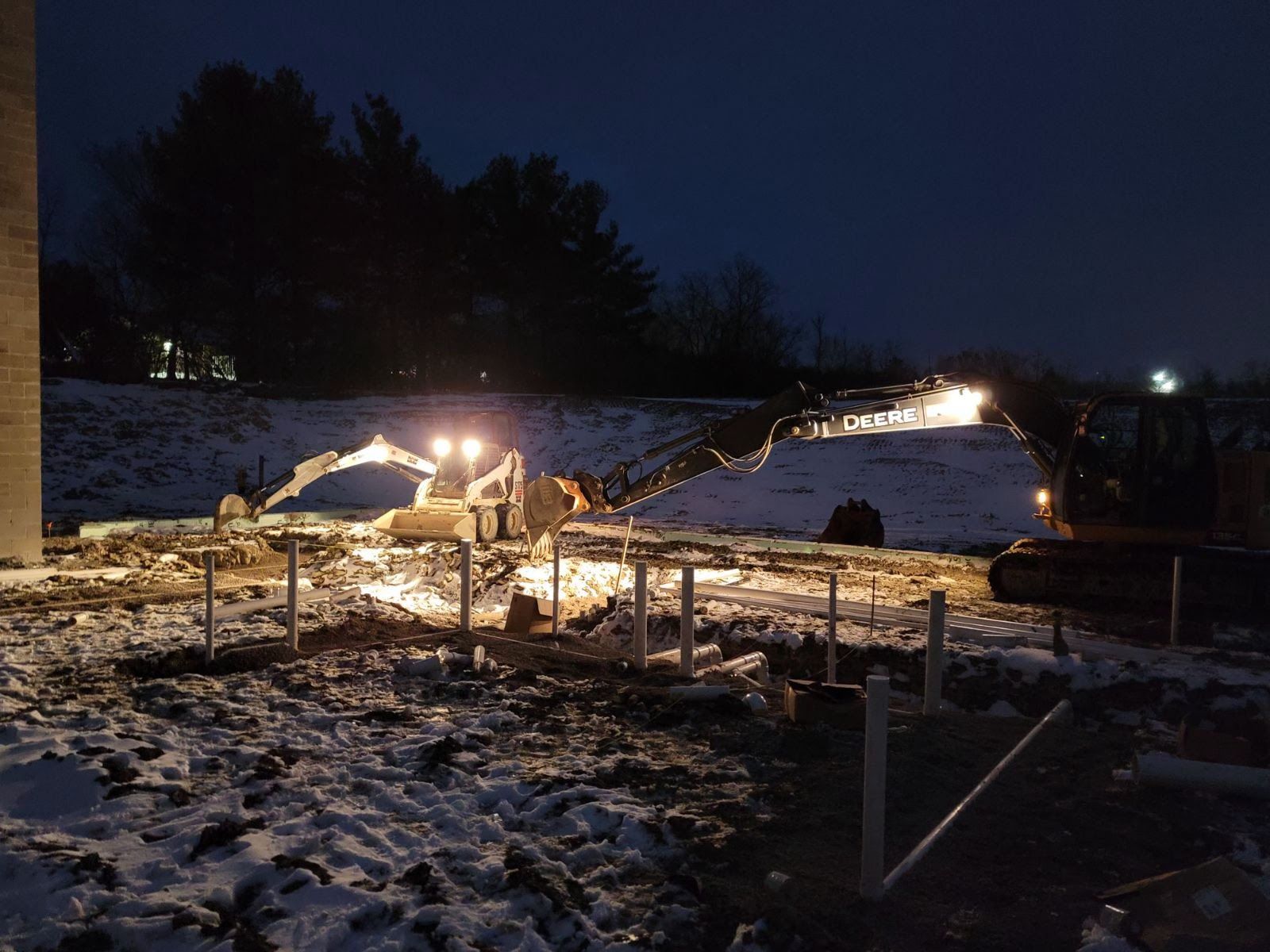 Construction site at night with excavator and lit work area, snowy ground.