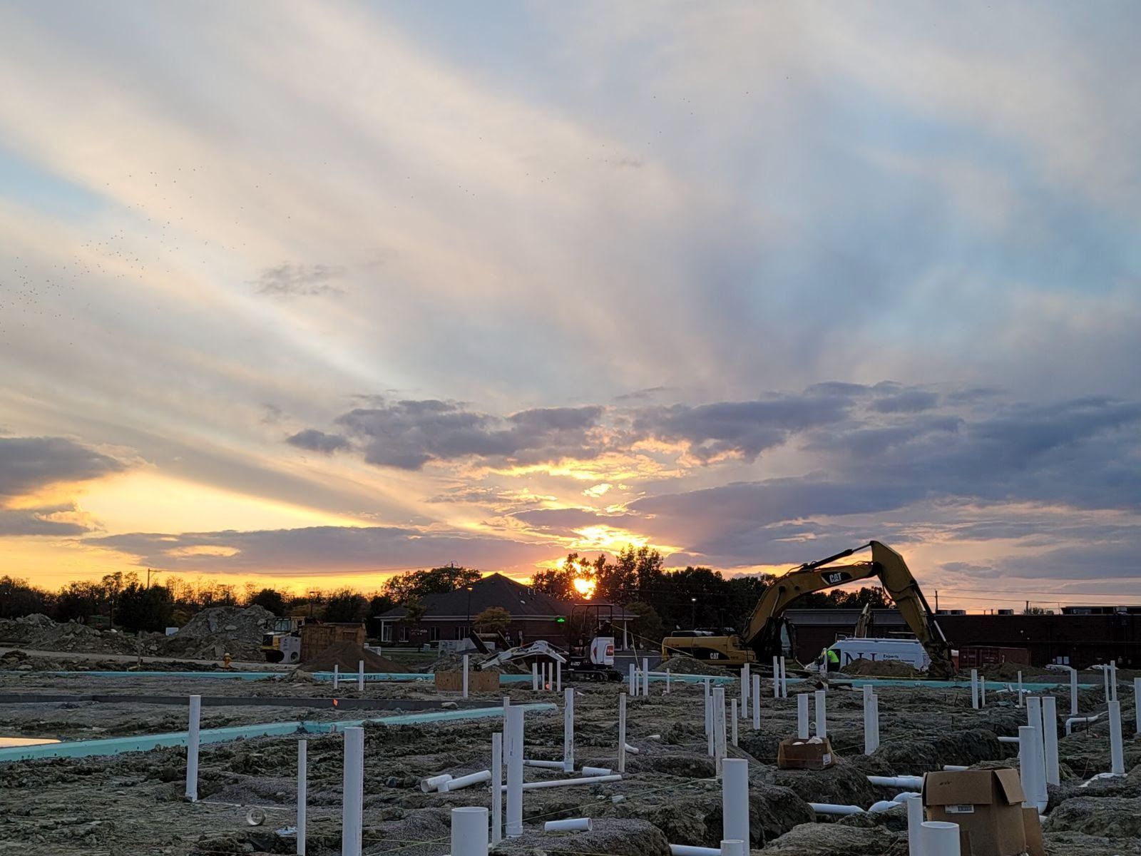 Construction site at sunset with excavator, clouds, and white pipes.
