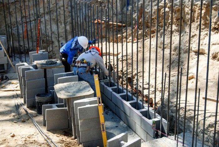 Construction workers building a concrete block wall, using levels and rebar in an outdoor setting.