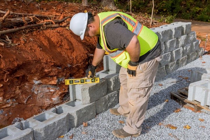 Construction worker using a level to check retaining wall bricks.  He's wearing a hard hat, vest, and gloves.