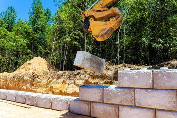 A yellow excavator placing a concrete block on a retaining wall during construction, outdoors in sunlight.