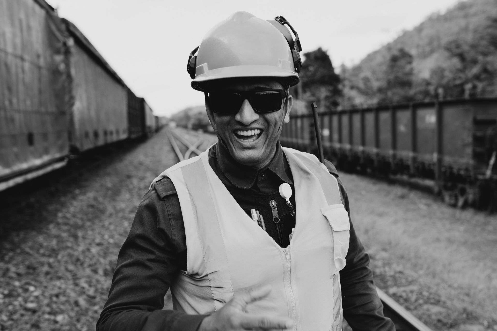 Smiling worker in safety gear stands on train tracks, near train cars.