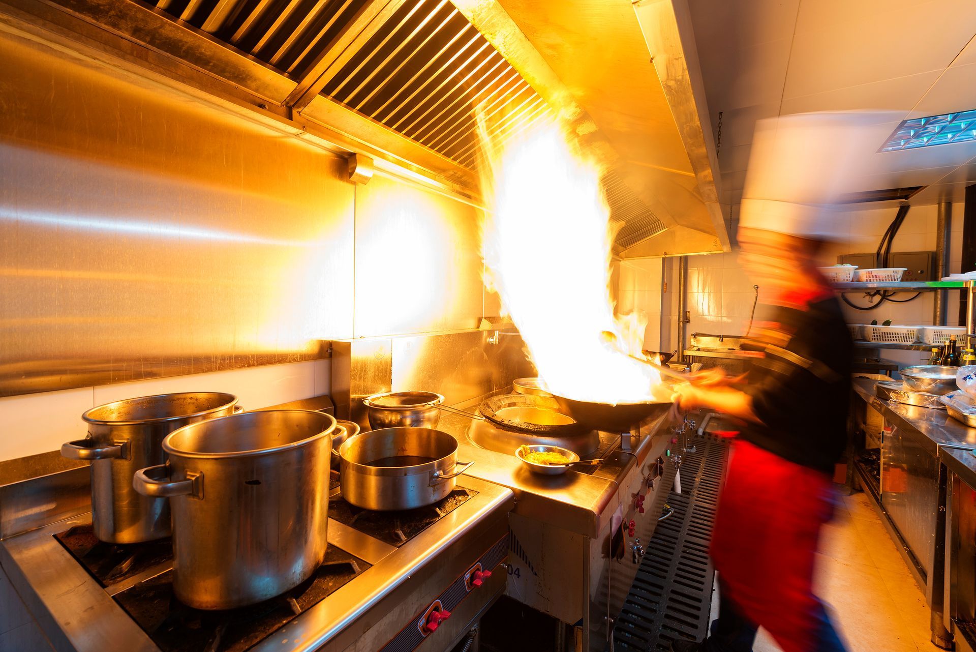 Kitchen stove with a wok on fire, cook blurred in the background. Stainless steel, bright flames.