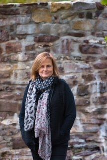 Woman in black coat and patterned scarf stands in front of a stone wall.