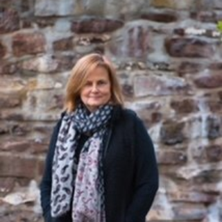 Woman in black coat and patterned scarf stands in front of a stone wall.