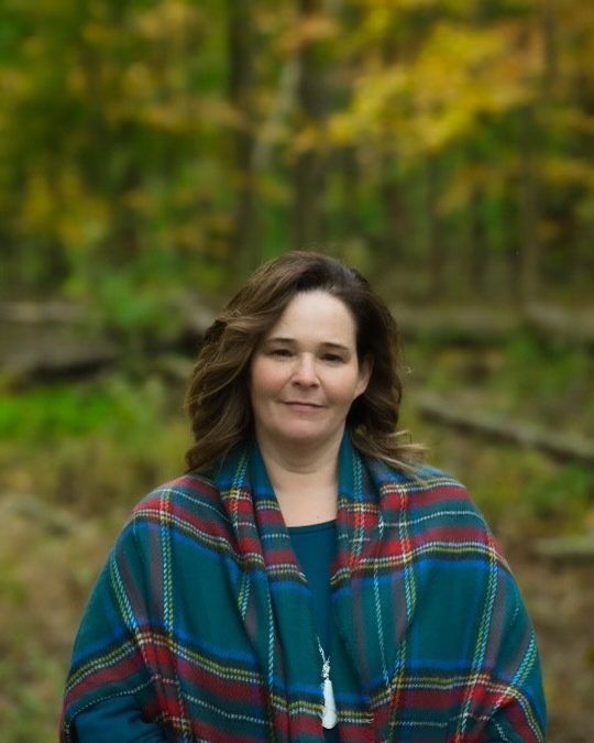 Woman in blue top and plaid shawl outdoors in front of fall foliage.