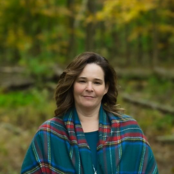 Woman in blue top and plaid shawl outdoors in front of fall foliage.