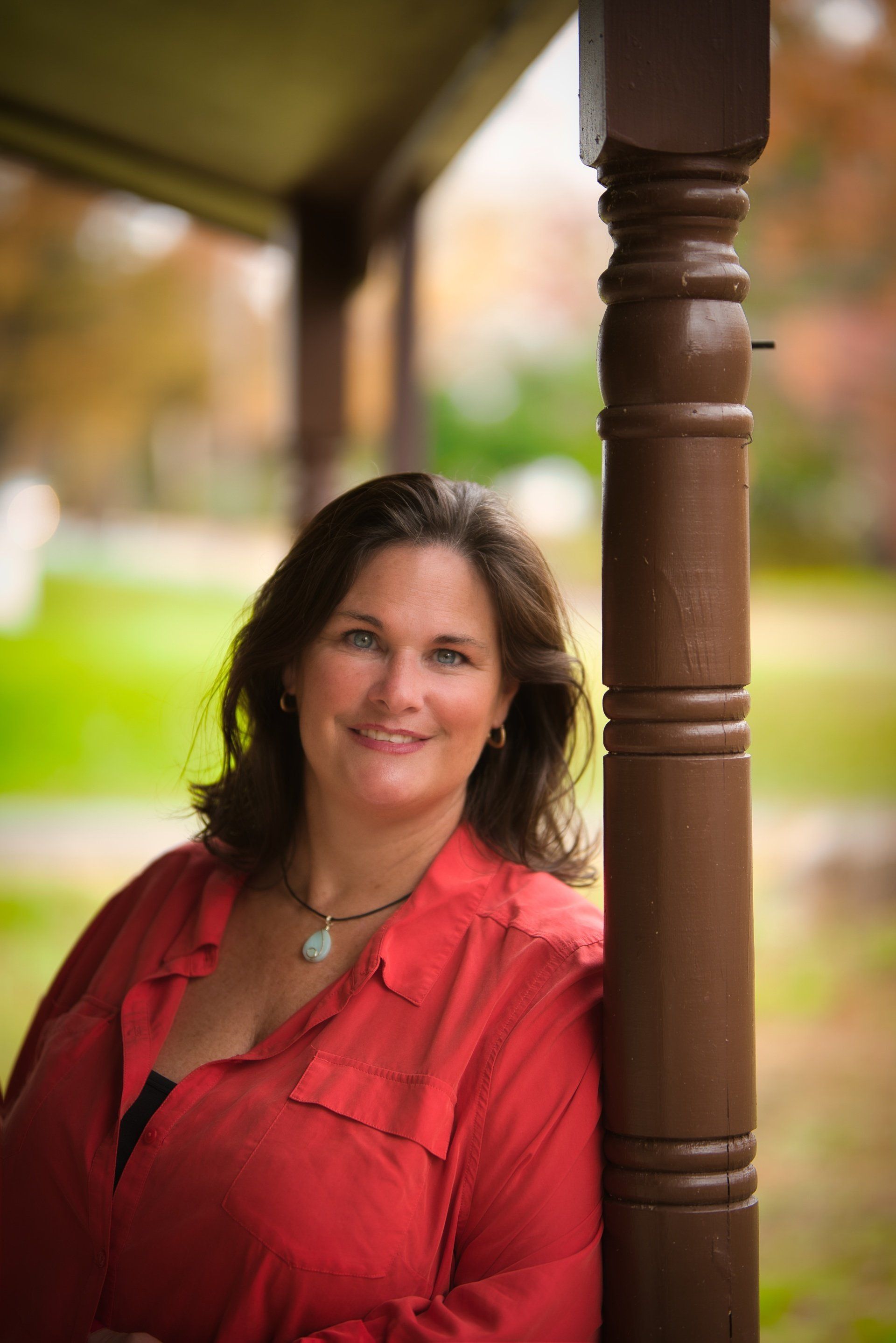 Woman in a red shirt smiles, leaning against a brown porch pillar, outdoors with blurred fall foliage.
