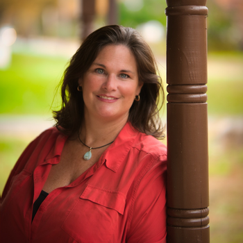 Woman in a red shirt smiles, leaning against a brown porch pillar, outdoors with blurred fall foliage.