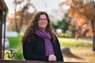 Woman with curly hair and glasses wearing a purple scarf and black sweater, smiles in an outdoor setting with trees.