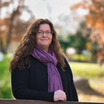 Woman with curly hair and glasses wearing a purple scarf and black sweater, smiles in an outdoor setting with trees.