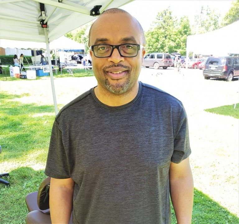 Man wearing glasses and a gray shirt smiles outdoors with a tent and vehicles in the background.