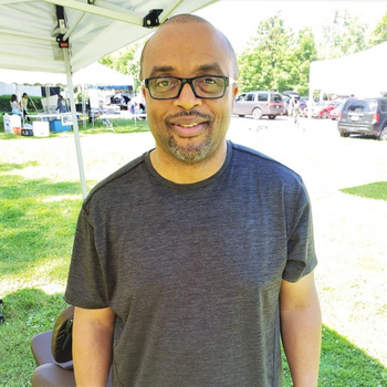 Man wearing glasses and a gray shirt smiles outdoors with a tent and vehicles in the background.