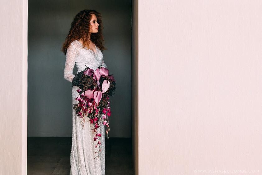 Bride in a white lace gown holding a large bouquet, standing in a doorway with a neutral backdrop.