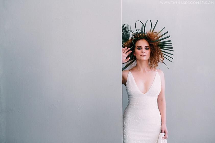 Woman in a white dress and elaborate headpiece poses next to a gray wall.