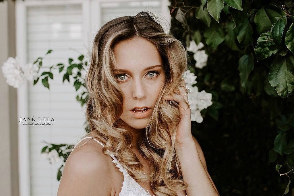 A woman with wavy blonde hair gazes at the camera, posing outdoors near white flowers and greenery.