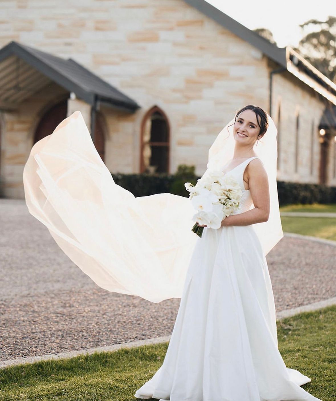 Bride in a white wedding dress and veil smiles, holding a bouquet, outside a stone church. The veil is blowing in the wind.