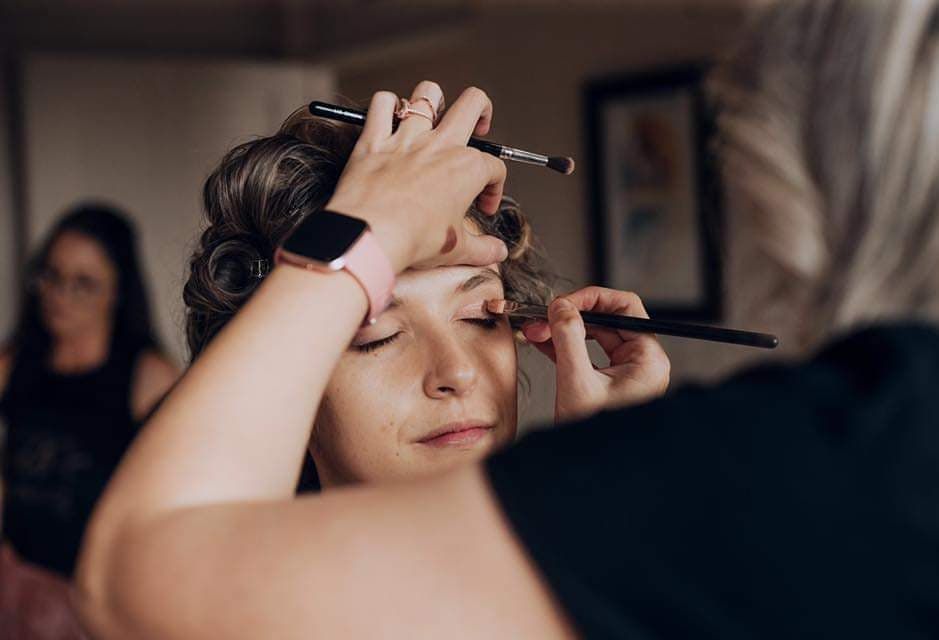 A makeup artist applies eyeshadow to a woman with curlers in her hair, inside a room. A second woman is in the background.