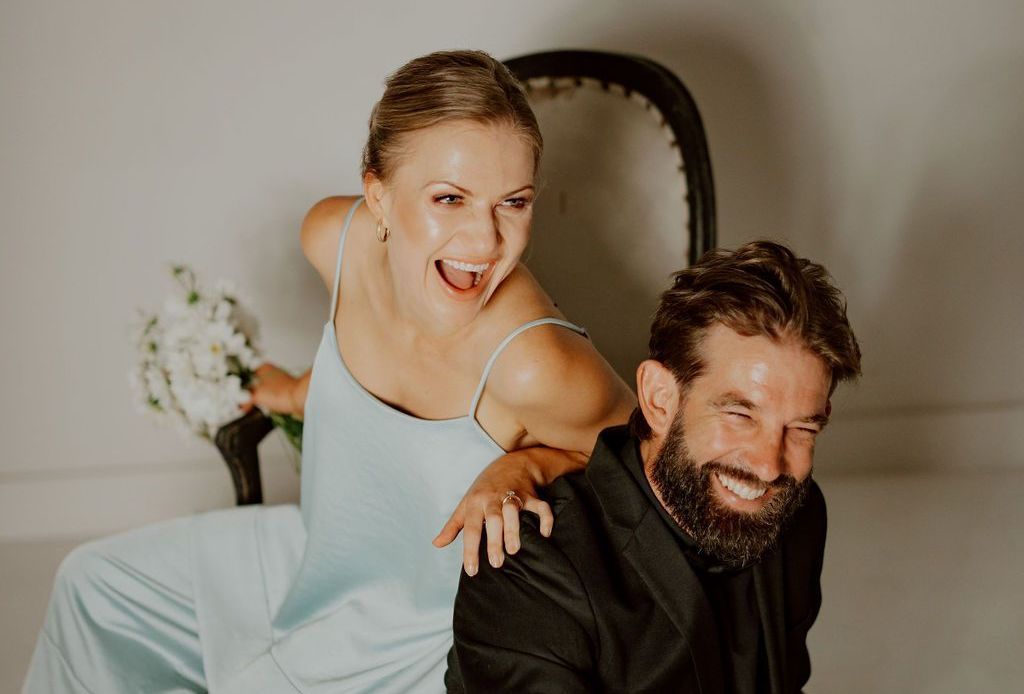 A joyful couple, woman in blue dress leaning over man's shoulder, both smiling. They're posing near a black chair.