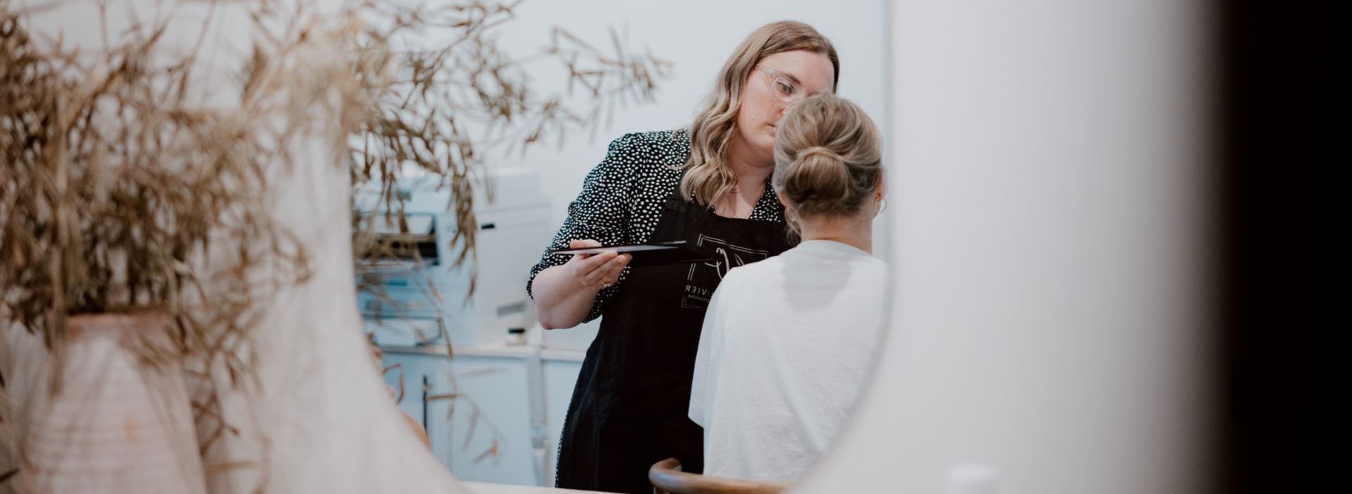 A woman in a patterned top and glasses consults with another woman with her hair up, in front of a mirror.