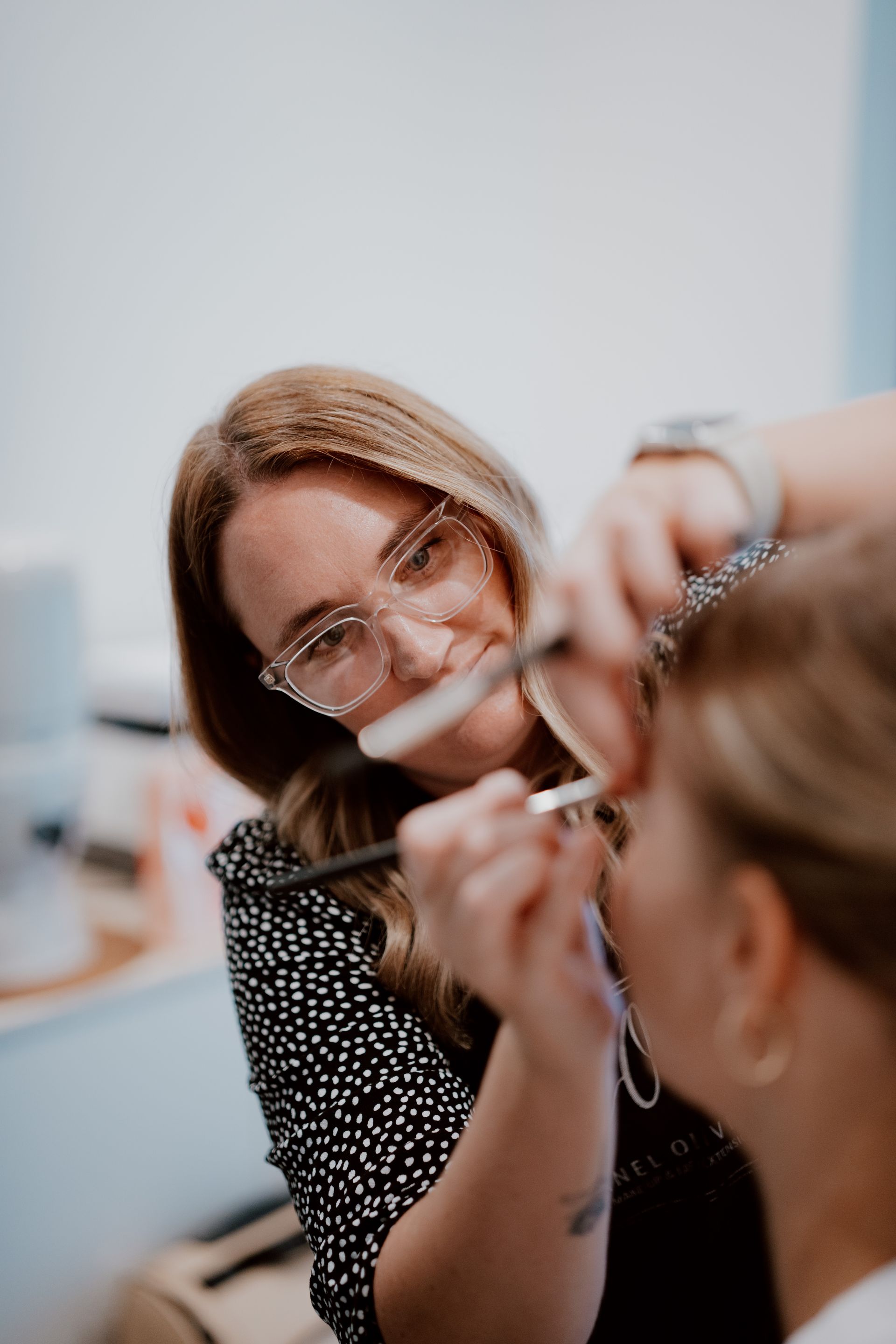 A makeup artist applies eyeshadow to a client's eye. The artist wears glasses and a black polka-dot shirt.