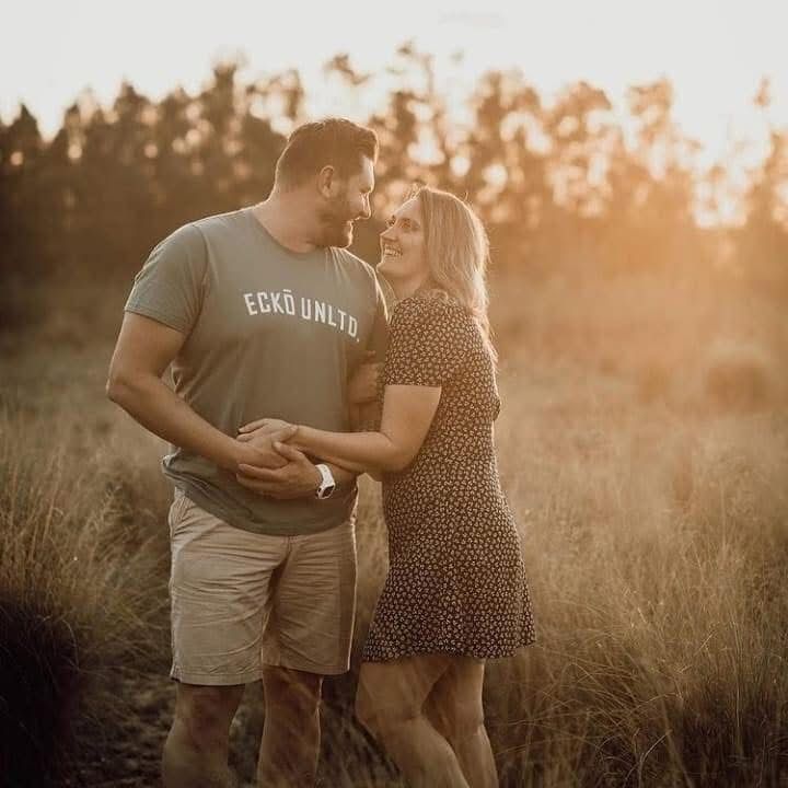 Couple holding hands and gazing at each other in a field at sunset. Man in green shirt, khaki shorts; woman in floral dress.