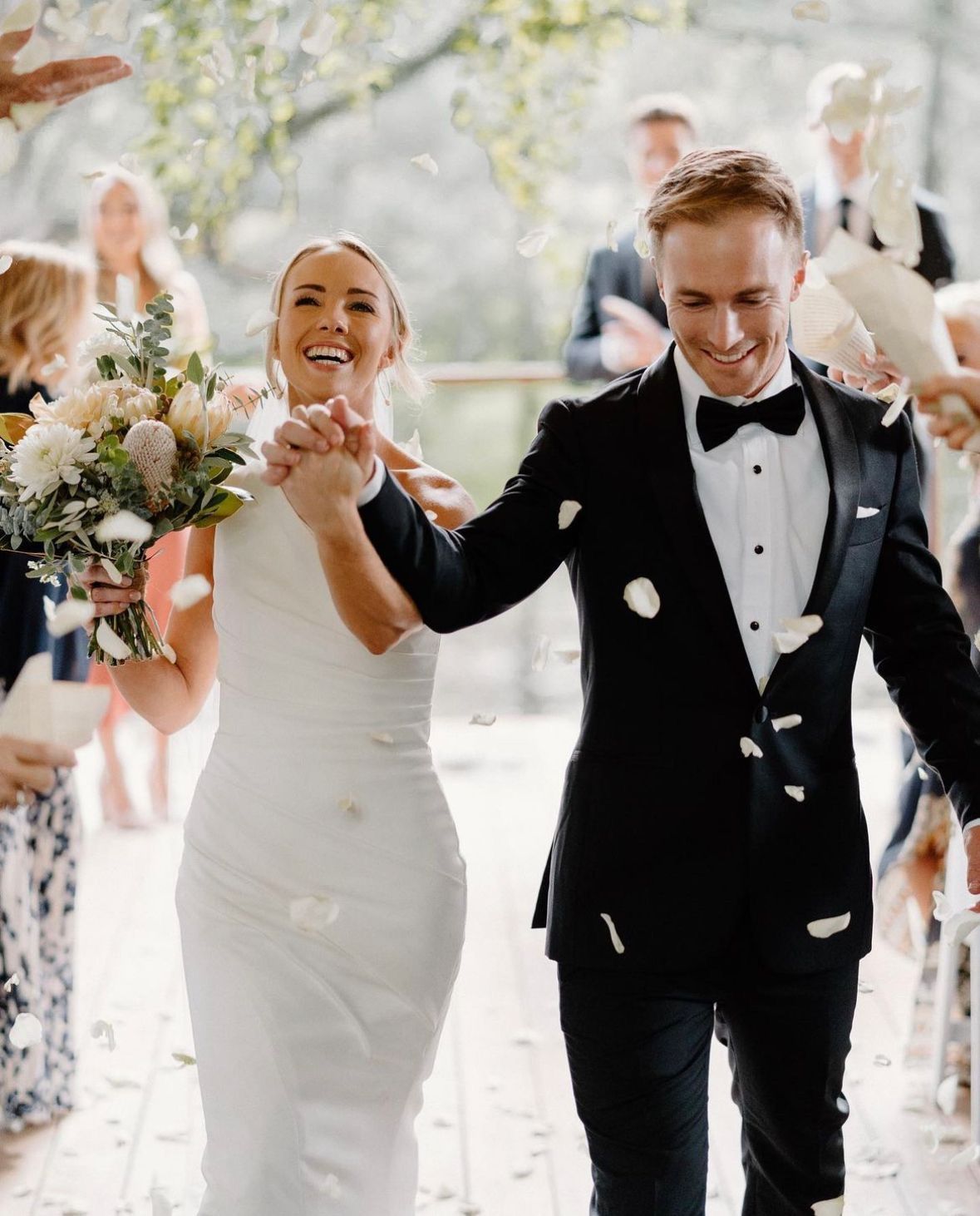 Newlyweds holding hands, smiling, and walking down an aisle as white flower petals are thrown. 
