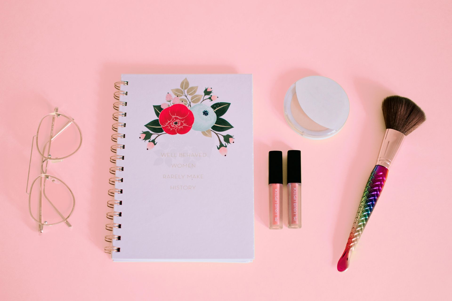A notebook with floral decorations, makeup, glasses, and a brush on a pink background.