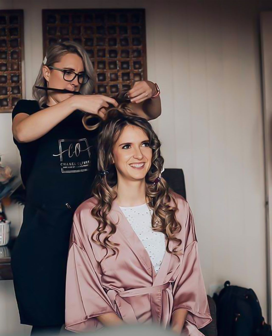 A hairstylist with glasses styles a woman's long, curly hair. Both are inside, the woman wearing a pink robe.
